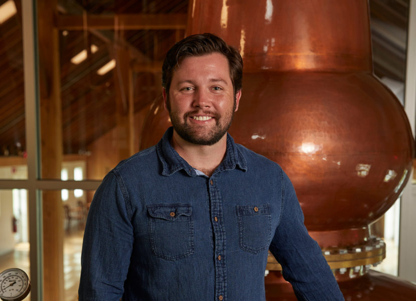 Cole Peck, Distiller, Tenmile Distillery Man in a dark denim button down leaning against a large whiskey still.