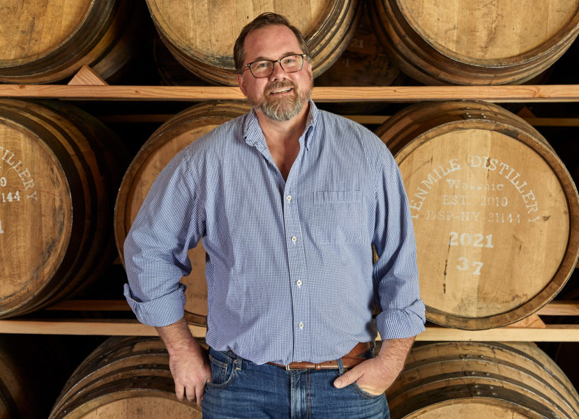 Man in a blue button down leaning against whiskey barrels.