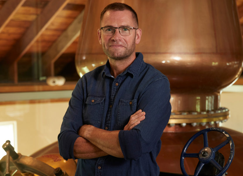 Shane Fraser, Master Distiller, Tenmile Distillery Man in a dark denim button down leaning against a large whiskey still.