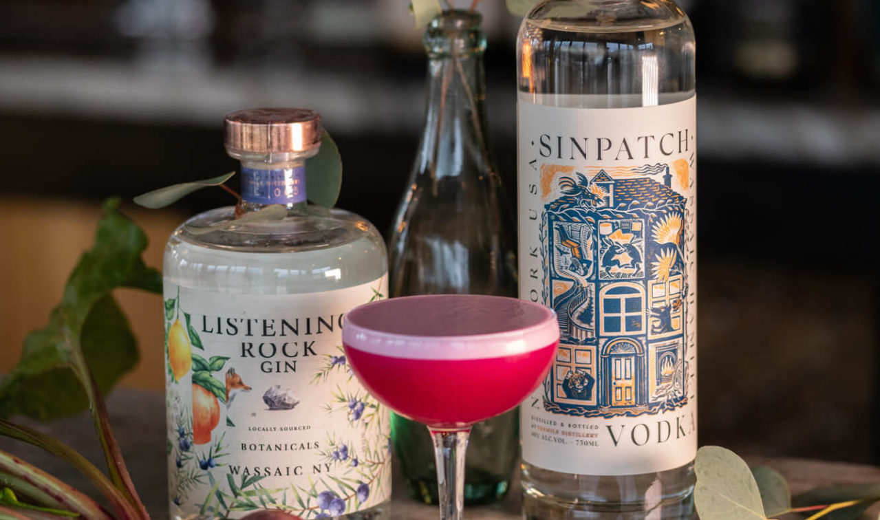 Dark pink cocktail in a glass on a bar next two two bottles of liquor.